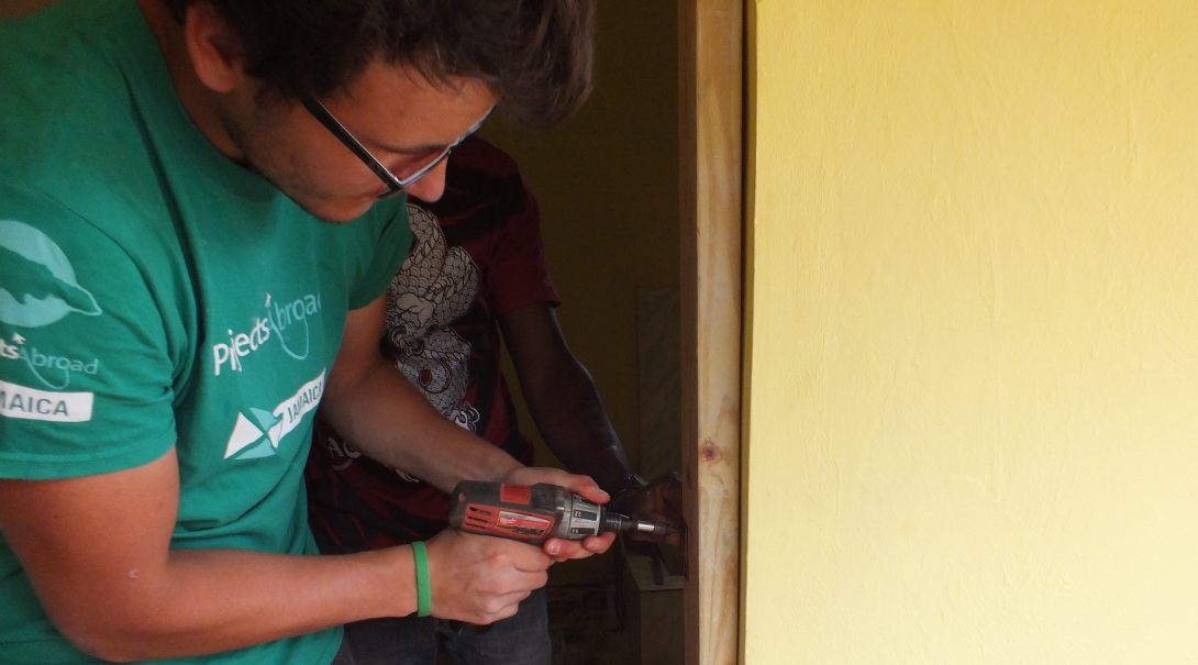 A Projects Abroad volunteer working with a group attaches a door to a door frame as part of building volunteer work in Jamaica. A volunteer helps with building a bathroom as part of construction volunteering in Jamaica for a group.